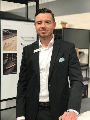 Geschäftsmann im dunklen Anzug mit Namensschild steht lächelnd an einem Messestand vor einer Präsentation moderner Griff-Designs.