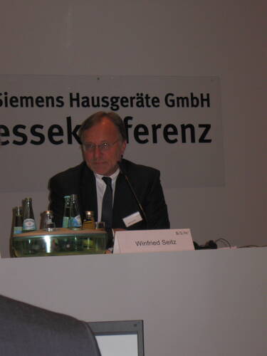 Manager spricht bei einer Pressekonferenz der Siemens Hausgeräte GmbH hinter einem Rednerpult mit Namensschild und Getränkeflaschen