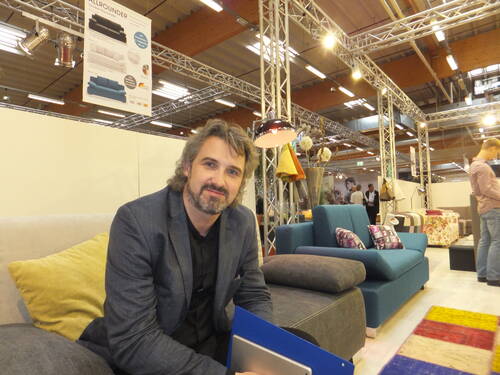 Mittelalterlicher Mann mit Bart sitzt lächelnd auf einem Sofa in einer modern beleuchteten Möbelmesse und hält eine blaue Mappe in den Händen.