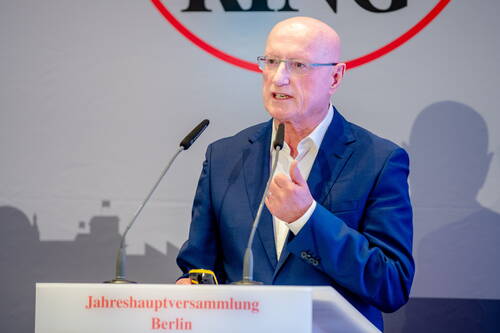 Älterer Mann im blauen Anzug spricht engagiert am Rednerpult mit zwei Mikrofonen während einer Jahreshauptversammlung in Berlin.
