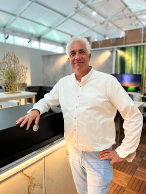 Älterer Mann mit weißem Haar und weißem Hemd lehnt lächelnd an einem modernen Grill in einer hellen Ausstellungshalle