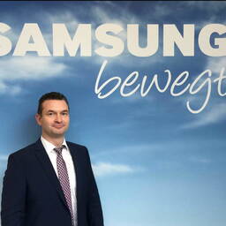 Geschäftsmann im Anzug steht vor einer blauen Wand mit großem Samsung-Schriftzug und dem Wort „bewegt“ in geschwungener Schrift.