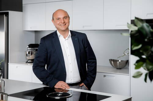 Lächelnder Geschäftsmann im Anzug steht entspannt an einer modernen weißen Kücheninsel mit Induktionskochfeld