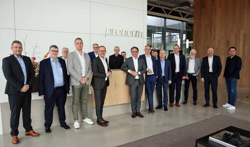 Gruppe von fünfzehn Männern in Anzügen und Businesskleidung posiert im modernen Eingangsbereich eines Unternehmens vor einem Tresen mit dem Schriftzug „pronorm“