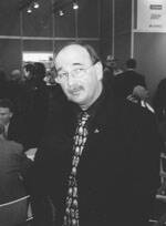 Mittleren Alters stehender Mann mit Brille und Schnurrbart in Anzug und gemusterter Krawatte auf einer geschäftigen Messe oder Konferenz in Schwarz-Weiß-Aufnahme