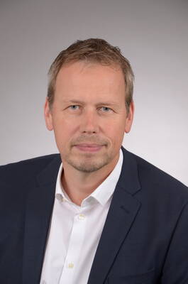 Mittelalterlicher Mann mit kurzem blondem Haar und Bartansatz im dunklen Anzug und weißem Hemd blickt ruhig in die Kamera vor neutral grauem Hintergrund