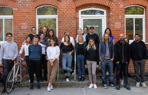 Große, lächelnde Bürogruppe steht lässig vor einem Backsteingebäude mit der Eingangstür zum KptnCook-Büro und posiert für ein Teamfoto.