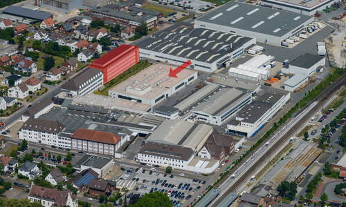 Luftaufnahme eines großen Industrie- und Bürokomplexes mit markiertem Flachdachgebäude im Zentrum und rot hervorgehobener mehrstöckiger Längsbebauung am Rand eines Wohngebiets neben Bahngleisen.