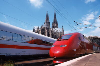 Roter Thalys-Hochgeschwindigkeitszug und weißer ICE stehen nebeneinander im Bahnhof, im Hintergrund ragen die Türme des Kölner Doms in den blauen Himmel.