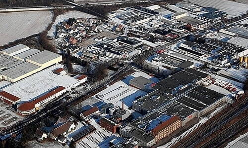 Luftaufnahme eines verschneiten Industriegebiets mit Lagerhallen, Produktionshallen und angrenzender Wohnsiedlung am Stadtrand
