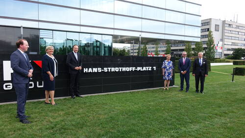 Sechs festlich gekleidete Personen stehen mit Abstand vor dem modernen Bürogebäude und dem schwarzen Namensschild „Hans-Strothoff-Platz 1“ auf einer gepflegten Rasenfläche.