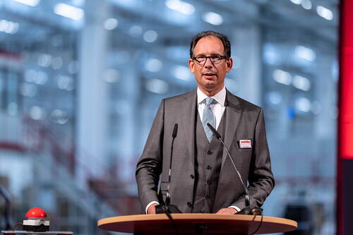Geschäftsmann im grauen Anzug spricht an einem Rednerpult mit zwei Mikrofonen in einer modernen, hellen Industriehalle.