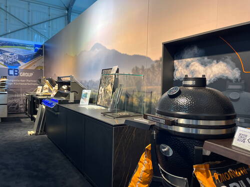 Rauchender Keramikgrill an einem modernen Messestand mit Outdoor-Küchenmodulen und beleuchteten Gasgrills vor einer Landschaftsbild-Wand.