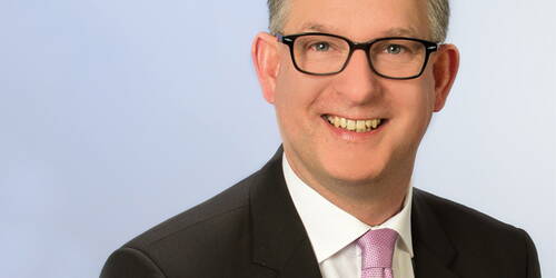 Lächelnder Mann mit Brille im dunklen Anzug und rosa Krawatte vor hellem Hintergrund