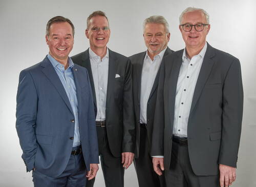 Vier lächelnde Geschäftsmänner mittleren bis höheren Alters posieren im Anzug nebeneinander vor neutralem, hellem Hintergrund.