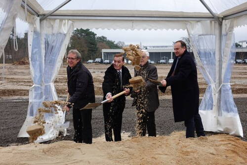 Vier Männer im Anzug heben bei einer feierlichen Grundsteinlegung unter einem weißen Zelt Sand mit Schaufeln aus einer vorbereiteten Baugrube.