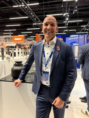 Lächelnder Mann im dunkelblauen Anzug mit Namensschild steht an einem Messestand in einer modernen Messehalle mit Design- und Küchenprodukten im Hintergrund.
