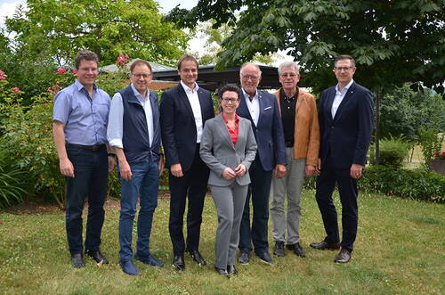 Sieben Personen in Business- und Freizeitkleidung stehen lächelnd nebeneinander auf einer Rasenfläche vor Bäumen und Sträuchern in einem Garten.