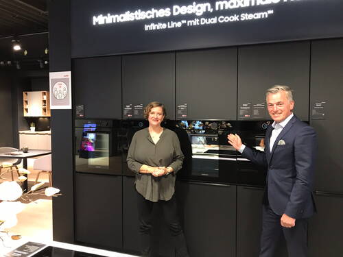 Zwei Personen präsentieren in einem modernen Küchenstudio eine schwarze Einbau-Backofenlinie mit der Aufschrift „Minimalistisches Design, maximale Funktion“ an einer dunklen Ausstellungswand.