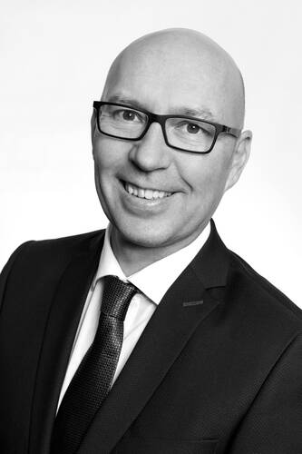 Lächelnder, glatzköpfiger Mann mit Brille im Anzug und Krawatte in einem professionellen Schwarz-Weiß-Porträt vor hellem Hintergrund