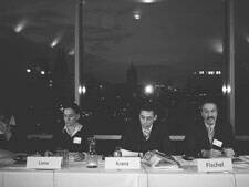 Drei Personen sitzen abends an einem Konferenztisch vor einer Fensterfront mit Stadtblick und lesen konzentriert Unterlagen.