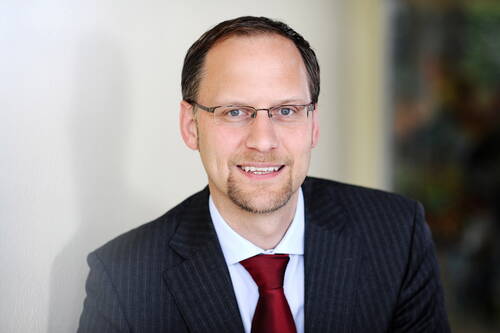 Lächelnder Geschäftsmann mit Brille im dunklen Nadelstreifenanzug und roter Krawatte vor neutralem Hintergrund