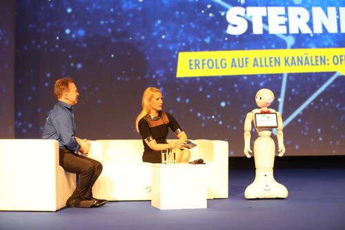 Moderatorin und Interviewpartner sitzen auf einer Bühne vor einer Sternenhintergrundprojektion, während ein humanoider Service-Roboter neben ihnen steht und am Gespräch teilnimmt.