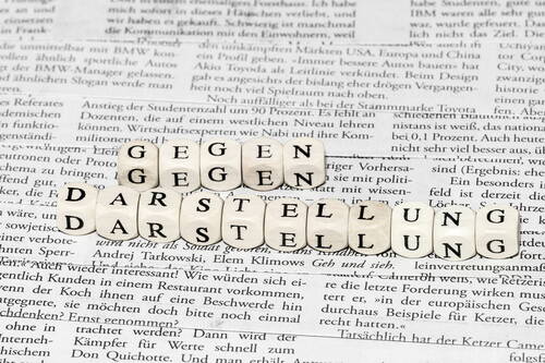 Holzplättchen mit den Wörtern „Gegen Darstellung“ liegen nebeneinander auf einer ausgebreiteten deutschsprachigen Zeitungsseite