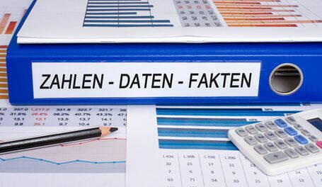 Blauer Ordner mit der Aufschrift „ZAHLEN – DATEN – FAKTEN“ liegt auf Finanzunterlagen mit Diagrammen, Taschenrechner und Bleistift