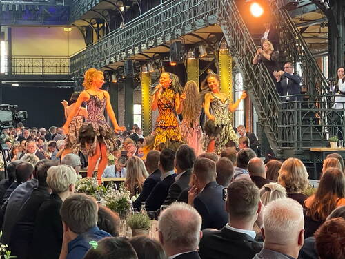 Tänzerinnen in farbenfrohen, federbesetzten Kleidern performen auf einer Bühne in einer historischen Industriehalle vor einem festlich gekleideten Publikum.