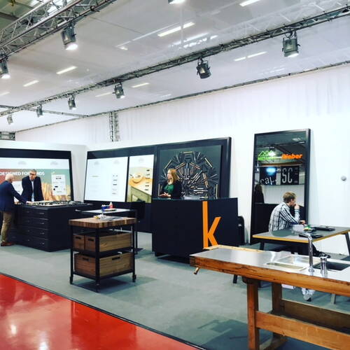 Moderner Messestand mit Küchenauszügen und Arbeitsplatten, an dem zwei Männer Produkte begutachten, eine Frau hinter einem Informationscounter steht und ein weiterer Mann an einem Tisch arbeitet.