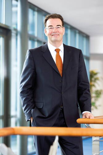 Geschäftsmann im dunklen Anzug mit orangefarbener Krawatte lehnt lächelnd an einem Handlauf in einem modernen, lichtdurchfluteten Bürogebäude.