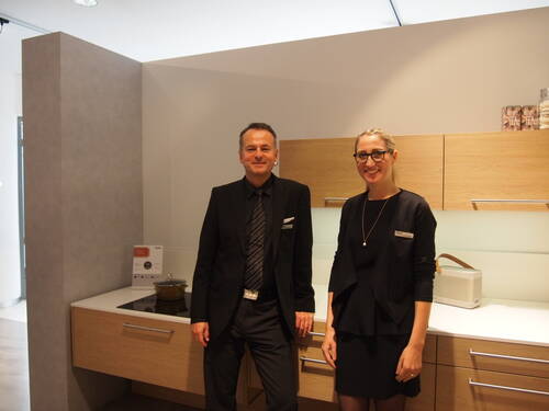 Zwei lächelnde Mitarbeitende in dunkler Businesskleidung stehen in einer modernen Ausstellungsküche mit hellen Holzmöbeln und weißer Arbeitsplatte.