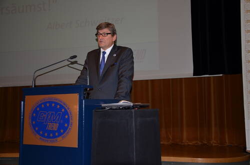 Mann im Anzug hält eine Rede an einem Rednerpult in einem Konferenzsaal vor einer Leinwand mit Zitat von Albert Schweitzer