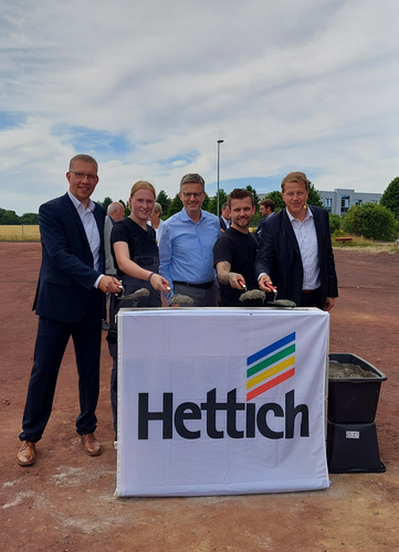 Fünf Personen halten Maurerkellen mit frischem Beton über einem mit Hettich-Logo versehenen Tisch und posieren bei einem feierlichen Spatenstich auf einem offenen Firmengelände.