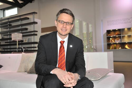 Geschäftsmann im Anzug mit rot gestreifter Krawatte sitzt lächelnd auf einem weißen Sofa in modern eingerichteter Ausstellungshalle