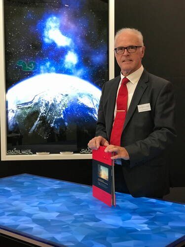 Älterer Mann im Anzug mit roter Krawatte steht an einem leuchtenden, blauen Präsentationstisch und hält eine rote Informationsmappe vor einem großformatigen Weltraum- und Erdbildschirm.