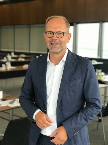 Mittelälterer Mann mit Brille und blauem Anzug steht lächelnd in einem modernen Konferenzraum mit leeren Tischen im Hintergrund