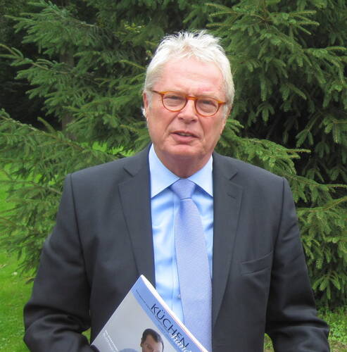 Älterer Mann im Anzug mit hellblauer Krawatte hält eine Zeitschrift in der Hand und steht vor einer grünen Tanne im Freien