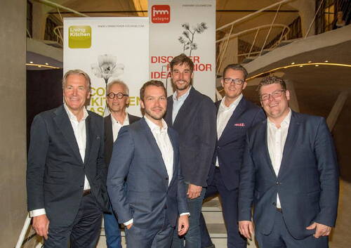 Sechs Männer in Anzügen stehen lächelnd vor Messeständen von LivingKitchen und imm cologne in einem modernen Ausstellungsraum