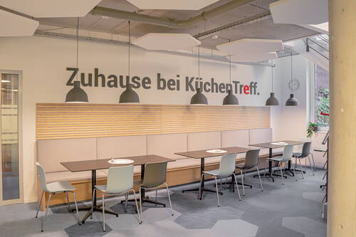 Modern gestalteter Pausenbereich mit langen Polsterbänken, mehreren Tischen und Hängelampen vor der Wandaufschrift „Zuhause bei KüchenTreff“