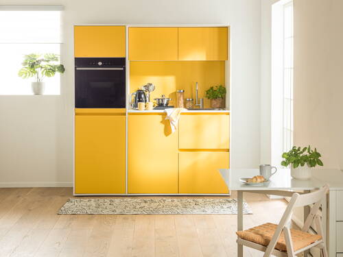 Helle, moderne Einbauküche mit gelben Fronten, integrierter Kaffeemaschine und kleinem Esstisch mit Croissant und Kräutertopf im Sonnenlicht