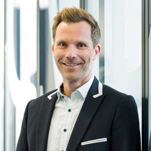 Lächelnder Mann mittleren Alters im Anzug mit weißem Hemd steht in einem modernen, hellen Büroflur und blickt direkt in die Kamera.