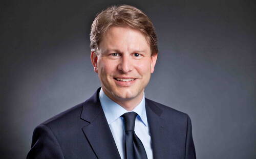 Lächelnder Mann im dunklen Anzug mit Krawatte vor neutral grauem Hintergrund