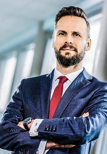 Selbstbewusster Geschäftsmann im blauen Anzug mit roter Krawatte steht mit verschränkten Armen in einem modernen, lichtdurchfluteten Büro.