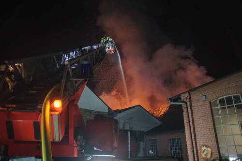 Feuerwehrleute löschen nachts von einer Drehleiter aus einen Großbrand in einem Fabrikgebäude, während Flammen und Funken in den Himmel schlagen