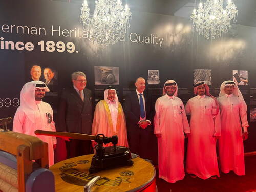 Delegation in traditionellen arabischen Gewändern und westlichen Anzügen posiert unter Kronleuchtern vor einer Ausstellungstafel zur deutschen Industriegeschichte seit 1899 mit historischen Maschinen im Vordergrund.