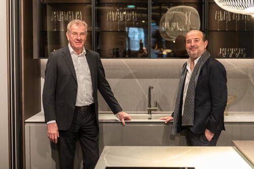 Zwei elegant gekleidete Männer stehen einander gegenüber in einer modernen Designküche mit Spülbecken, Glasvitrinen und stilvoller Beleuchtung.