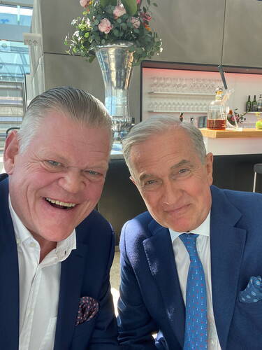 Zwei elegant gekleidete ältere Männer in blauen Anzügen lächeln in die Kamera, während sie in einer modernen Bar mit gläsernen Regalen und hoher Silbervase im Hintergrund zusammensitzen.