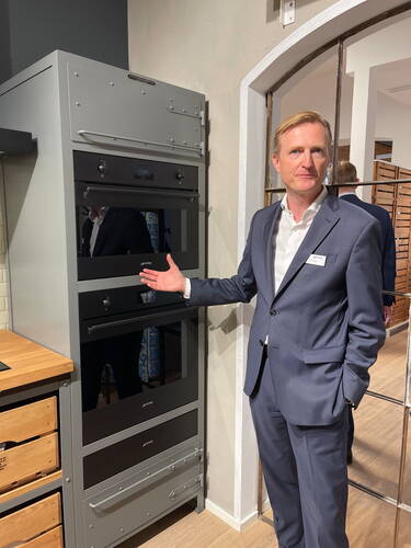 Mann im Anzug präsentiert in einer modernen Ausstellungsküche einen hohen, grauen Einbauschrank mit mehreren schwarzen Smeg-Backöfen und Stauraumfächern.